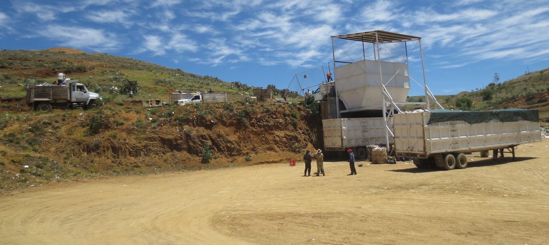 A large truck and two people stand near a white structure at a landfill or waste processing site on a sunny day. A pickup truck and more people are visible on the dirt hill in the background under a partly cloudy sky.