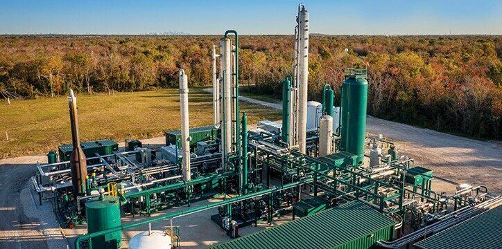 An industrial natural gas processing plant with tall metal structures, pipes, and green equipment, situated outdoors near a field and a forested area under a clear sky.