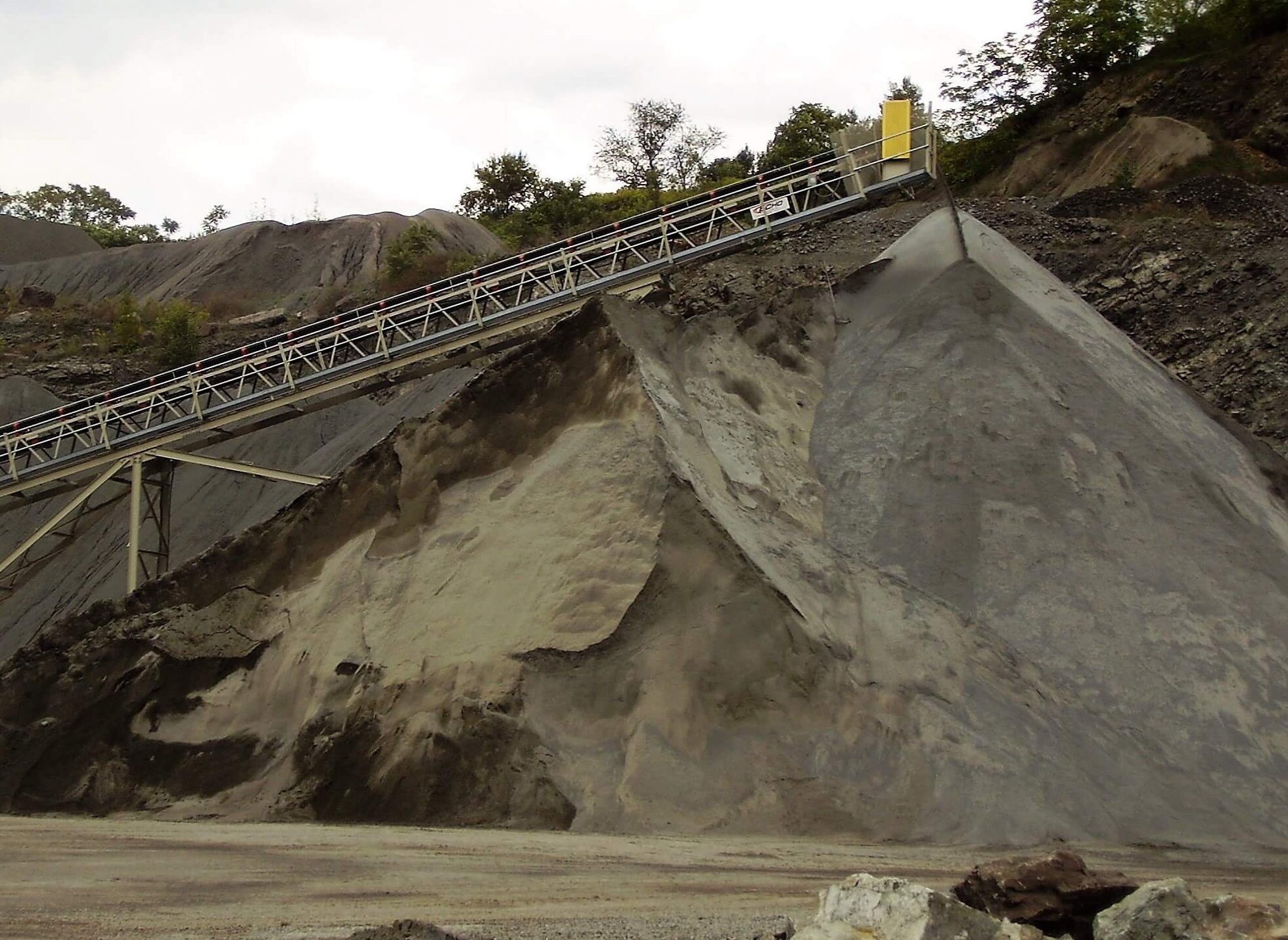A long conveyor belt deposits material onto a large, sloped pile of gravel and sand at an outdoor industrial site, with trees and rocky terrain in the background.