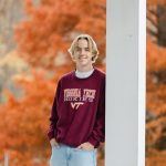 A young person with blond hair, wearing a maroon Virginia Tech Hokies sweatshirt and light jeans, stands outdoors by a white column with vibrant orange autumn foliage in the background.