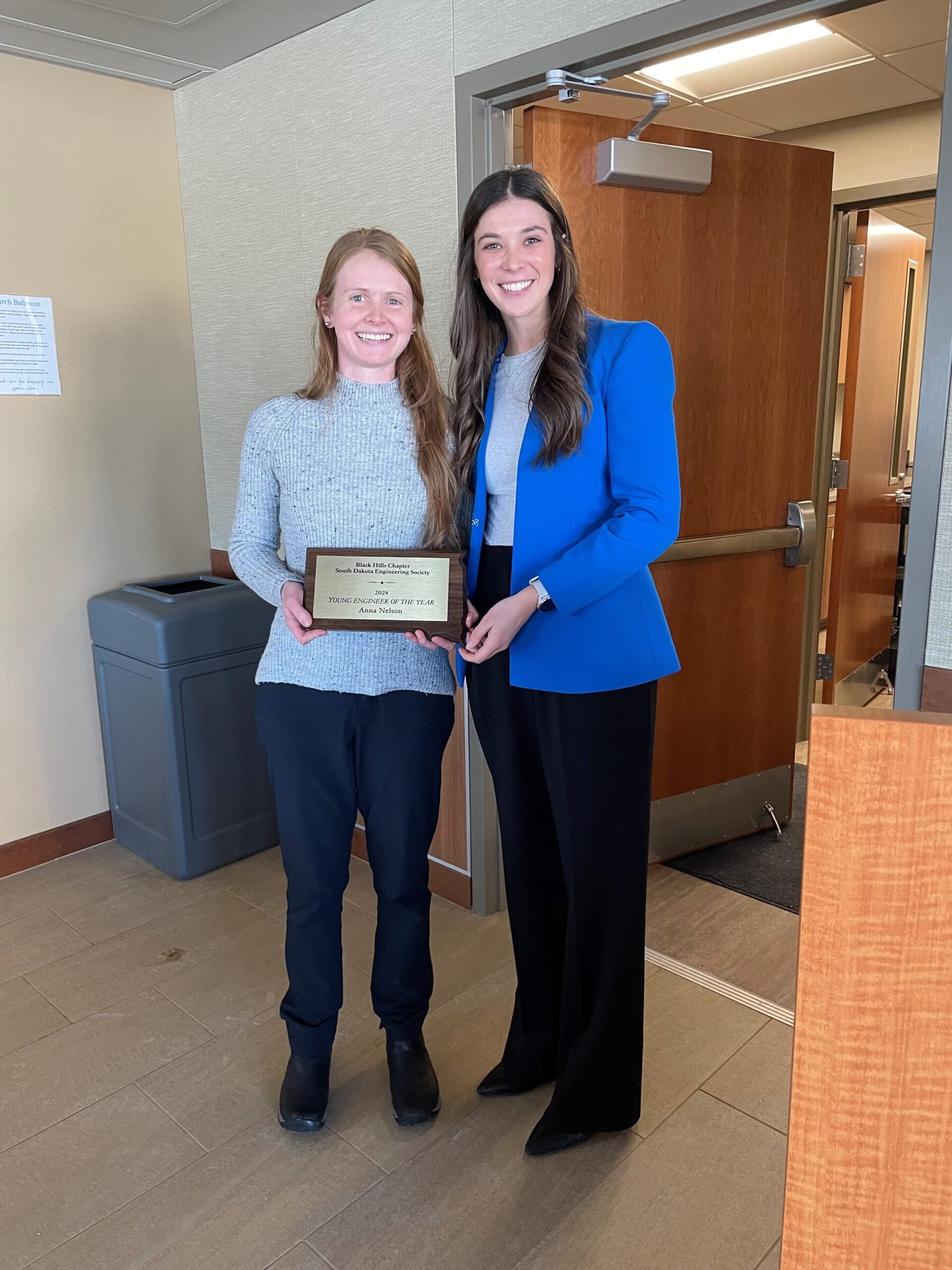 Two women stand indoors, smiling at the camera. One, in a gray sweater, holds a plaque with both hands. The other, in a blue blazer, stands beside her. They appear to be at an event or awards ceremony.
