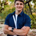 A young man with short brown hair stands outdoors, leaning against a tree with his arms crossed and smiling. He is wearing a navy blue short-sleeve shirt over a white t-shirt and a round-faced smartwatch.