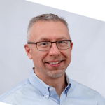 A smiling middle-aged man with short gray hair, glasses, and a light blue button-down shirt, posed in front of a plain white background. The photo is slightly rotated.