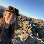 A young woman in a hat and sunglasses takes a selfie atop a rocky mountain peak with rugged cliffs and a clear blue sky in the background, bathed in golden sunlight.