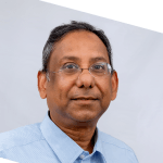 A middle-aged man with short dark hair, wearing glasses and a light blue collared shirt, smiles slightly while facing the camera against a plain light background. The image is tilted at an angle.
