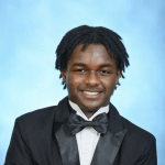 A young man with short dreadlocks smiles at the camera, wearing a black tuxedo, bow tie, and white shirt against a light blue studio background.