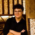 A young person with curly hair and glasses, wearing a black shirt, stands indoors with arms crossed, leaning against a wooden wall and smiling at the camera.