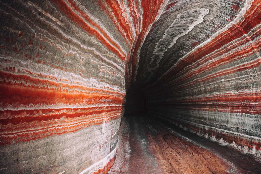 A tunnel with colorful, layered mineral stripes in red, white, and gray runs along curved walls and ceiling, creating a striking pattern as it recedes into darkness.