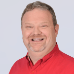 A smiling middle-aged man with short light brown hair and a goatee, wearing a bright red collared shirt, is posed in front of a plain light background.