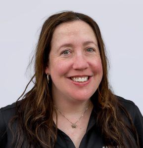 A woman with long brown hair wearing a black shirt, silver necklace, and hoop earrings smiles at the camera against a plain light background.