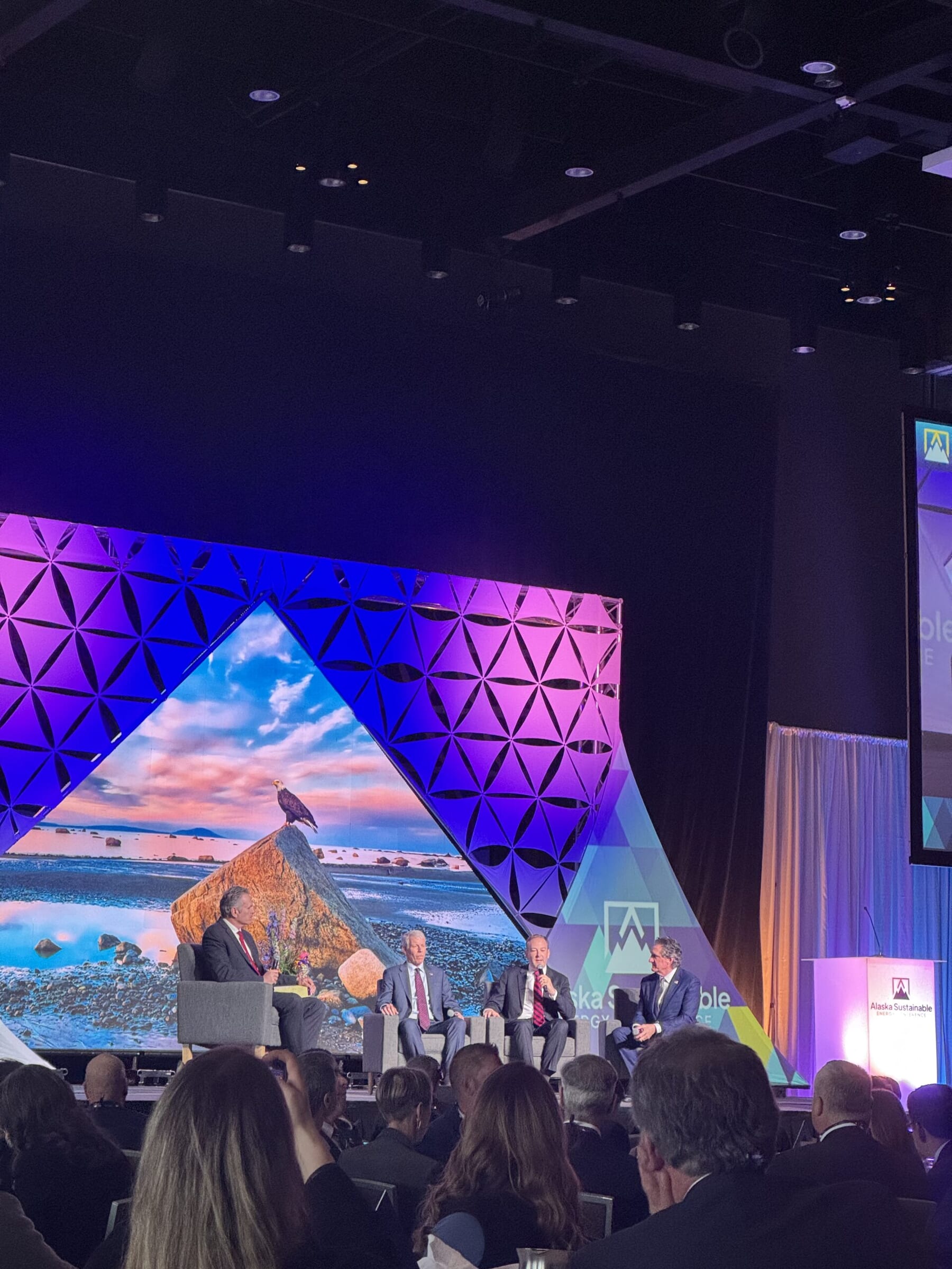 A conference panel on stage features four men in suits seated and speaking. Behind them is a large screen showing a scenic image of rocks and water, and geometric purple lighting decorates the stage. Audience members watch in the foreground.