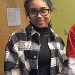 A young woman with glasses and curly hair wears a black and white plaid jacket over a black turtleneck. She is standing indoors, smiling slightly, with a green wall and wooden door behind her.