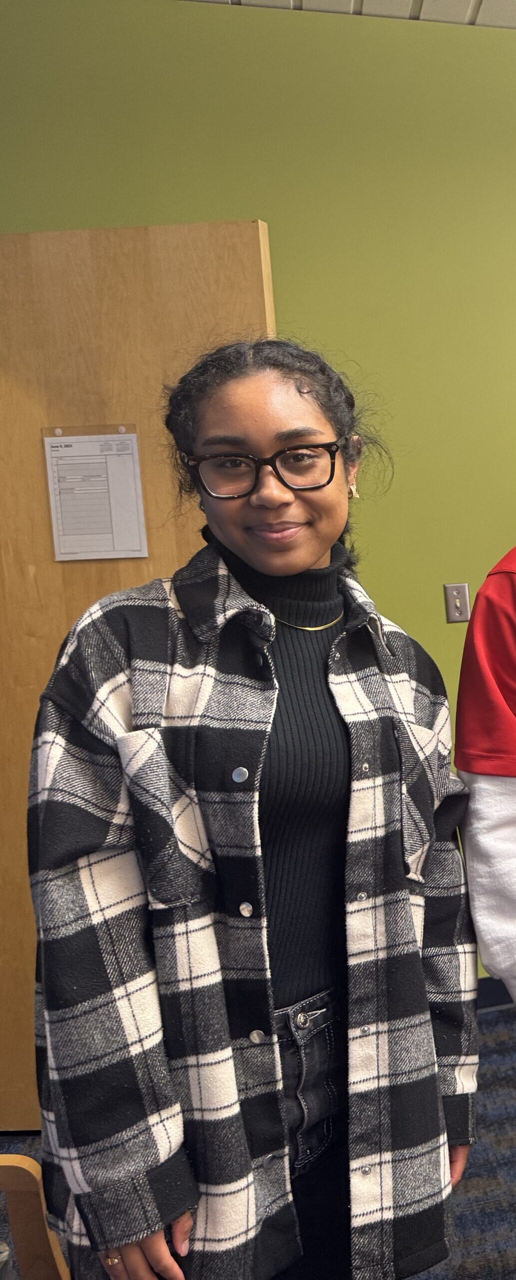 A young woman with glasses and curly hair wears a black and white plaid jacket over a black turtleneck. She is standing indoors, smiling slightly, with a green wall and wooden door behind her.