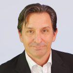 A middle-aged man with dark hair, wearing a black blazer and a white collared shirt, is looking at the camera against a plain light background.