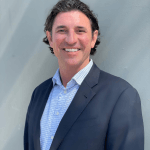 A man with wavy dark hair, wearing a navy blazer over a light blue checked shirt, smiles while standing in front of a light gray wall with soft shadows.