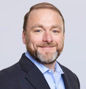 A man with light skin, short brown hair, and a trimmed beard smiles at the camera. He is wearing a dark blazer over a light blue collared shirt against a light gray background.