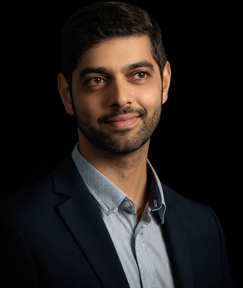 A man with short dark hair and a trimmed beard wearing a navy blazer and light blue shirt, smiling slightly and looking off to the side against a black background.