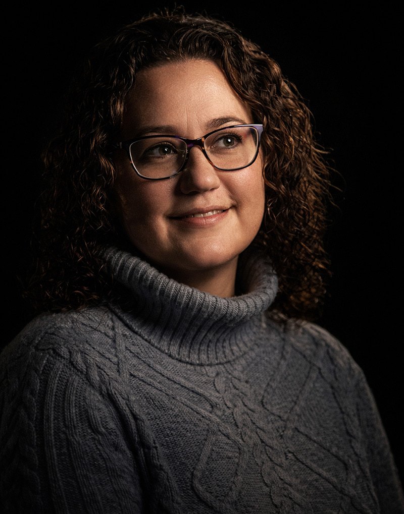 A woman with curly brown hair and glasses, wearing a gray cable-knit turtleneck sweater, smiles softly while looking slightly to the side against a dark background.
