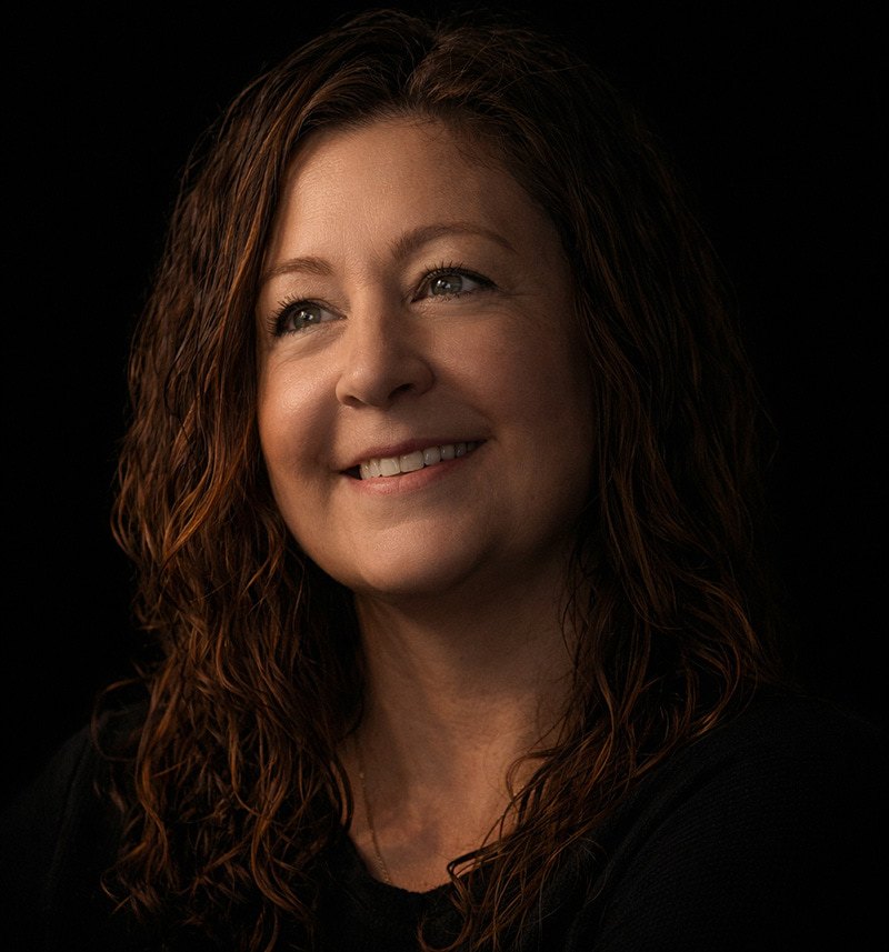A woman with long, wavy brown hair and a warm smile looks slightly to the side against a black background, wearing a dark top. The lighting is soft, highlighting her face.
