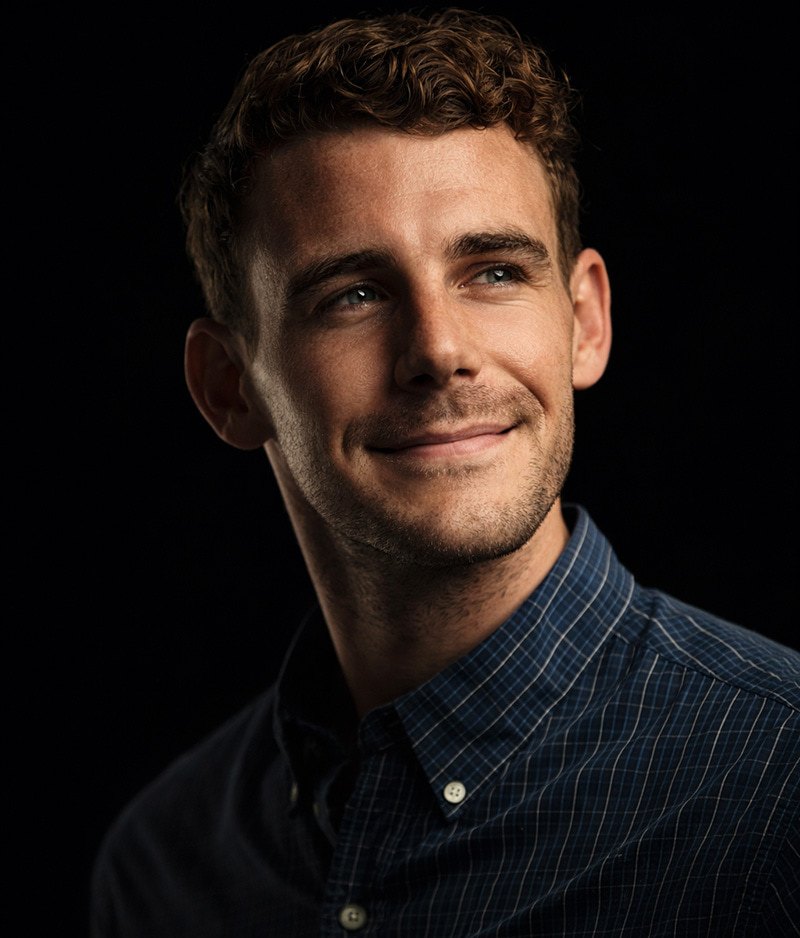 A young man with short, curly brown hair and light stubble wears a blue checkered shirt and smiles softly, looking slightly to the side against a dark background.
