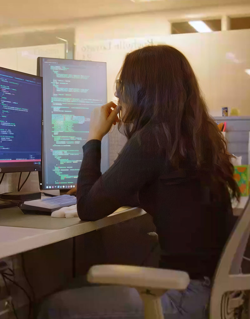 A woman with long brown hair sits at a desk, looking intently at a computer monitor filled with lines of code. She appears focused and thoughtful, using one hand to support her chin.