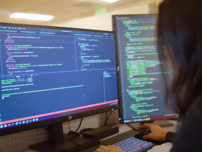 A person is working at a desk with two large monitors displaying code in a dark-themed text editor. The individual is typing on a keyboard, focusing on the screens filled with colorful programming syntax.