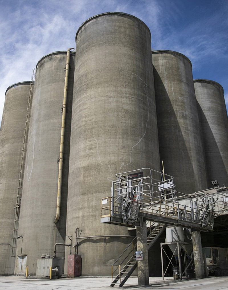 Five large, cylindrical concrete silos stand side by side, with industrial stairs, pipes, and platforms attached near the base. A blue sky with light clouds is in the background.