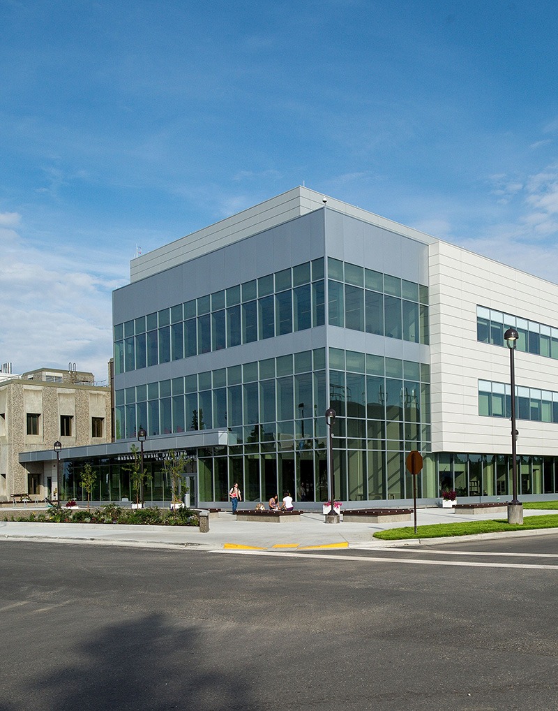 A modern three-story building with large glass windows and a gray and white exterior, surrounded by a sidewalk, lamp posts, and greenery beneath a blue sky.