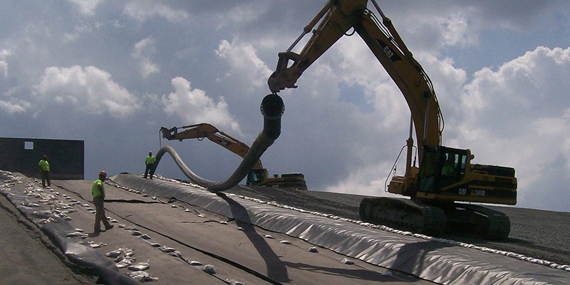 Four workers in safety gear supervise two large excavators as they lay geomembrane liner on a sloped surface under a cloudy sky. The excavators are positioning and unrolling the liner material.