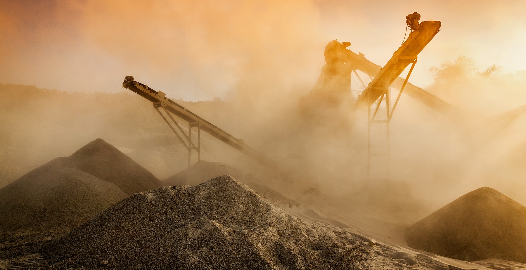 Two large industrial conveyor belts move sand or gravel at a quarry, surrounded by thick dust and piles of material under an orange, hazy sky.