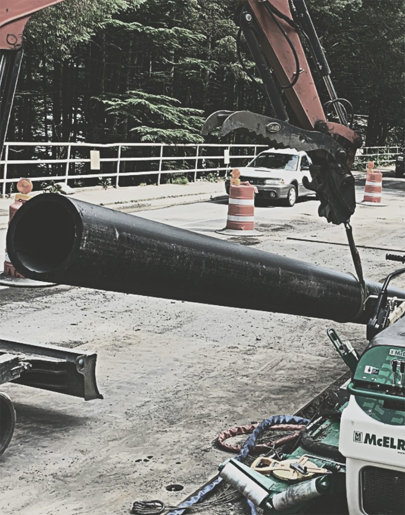 An excavator lifts a large black pipe at a construction site. Traffic cones, a parked car, and trees are visible in the background. Construction equipment and hoses are on the ground.
