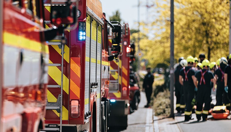 Several fire trucks with flashing lights are parked on a street while a group of firefighters in uniform and helmets stand together on the sidewalk, appearing to prepare for action.