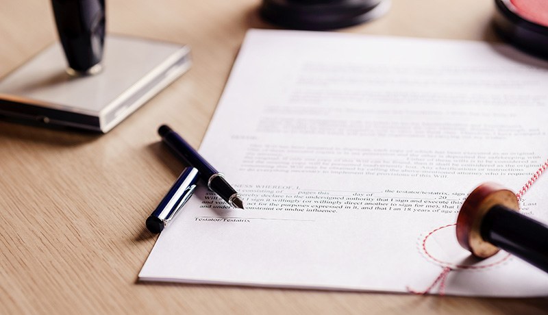 A close-up of a legal document on a desk, with a pen, an ink stamp, and a wax-sealed stamp nearby, suggesting a formal signing or notarization process.