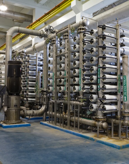 Industrial desalination equipment with rows of cylindrical white filtration membranes, pipes, and valves arranged vertically in a large facility with blue flooring and overhead ducts.