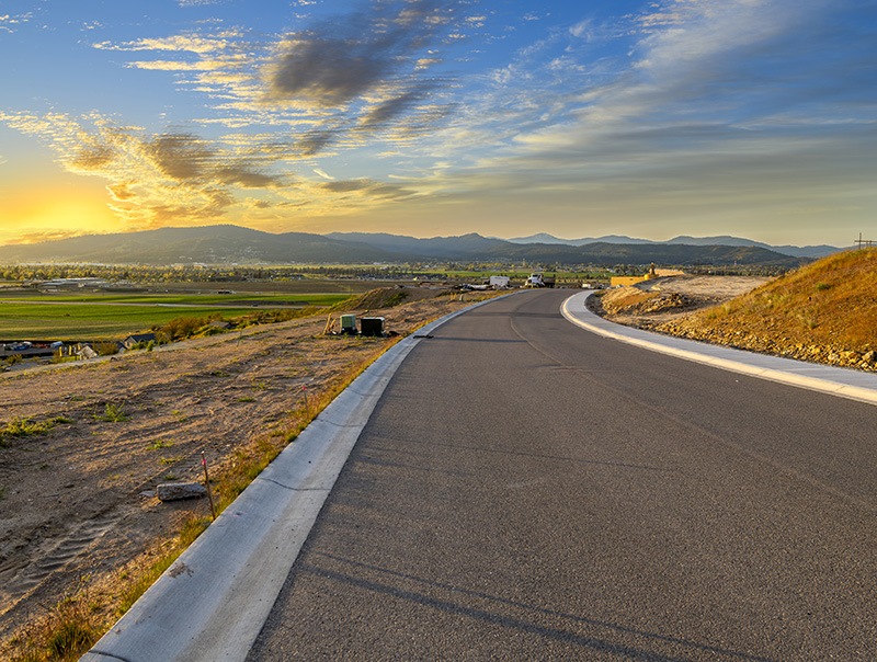 A freshly paved road curves through a rural landscape at sunset, with grassy fields and distant mountains under a sky with scattered clouds. Construction materials line the roadside.