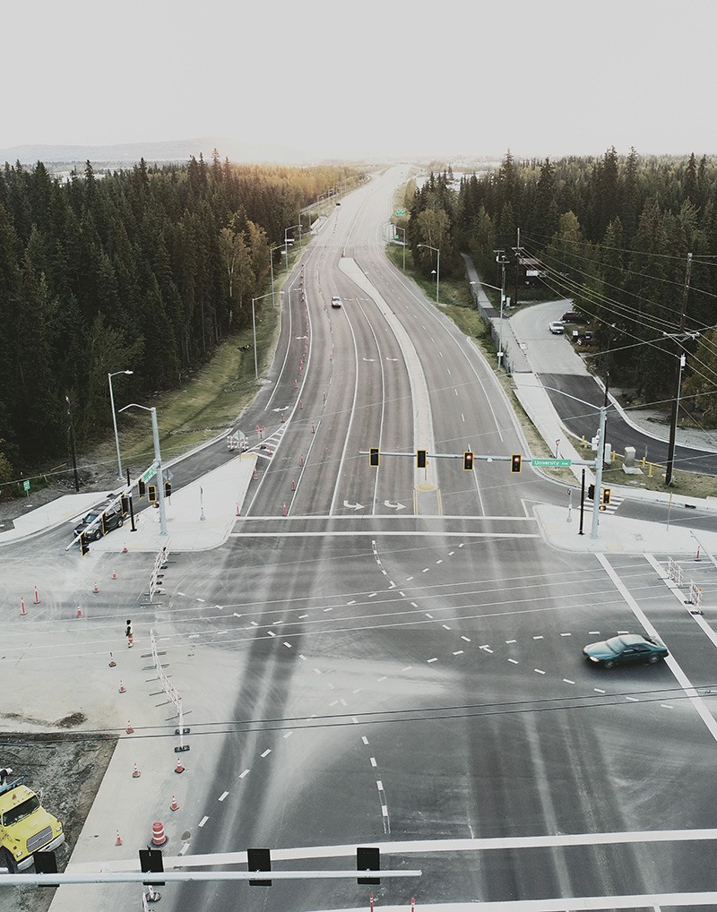 A wide, empty intersection with multiple lanes and traffic lights, surrounded by trees. A single car drives through, and another vehicle approaches, with few cars visible on the distant road. The sky is overcast.