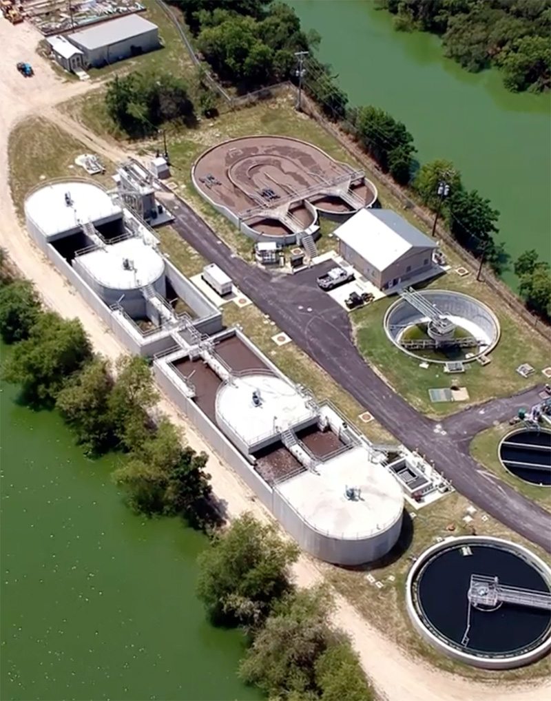 Aerial view of a water treatment facility with several large circular and rectangular tanks, a small building, paved roads, and surrounding greenery near a body of water.
