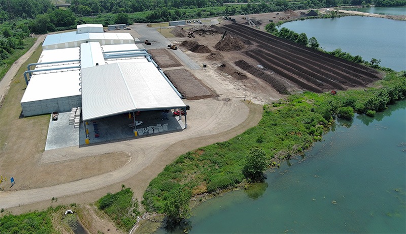 Aerial view of a large industrial facility near a body of water, with long rows of soil or compost piles, adjacent greenery, and several white-roofed buildings.