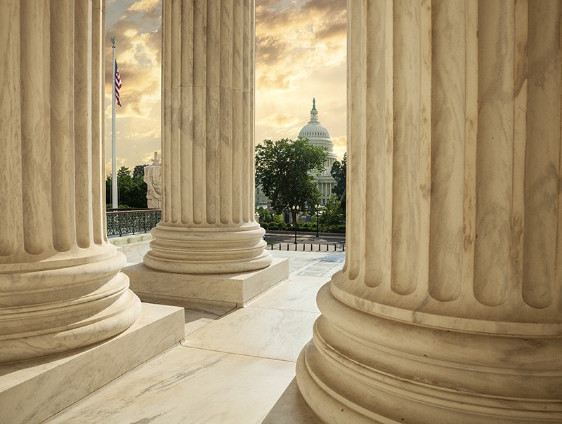 Sunlight streams through large marble columns with the US Capitol building visible in the distance, framed by the columns. An American flag waves to the side and trees line the open space between the structures.