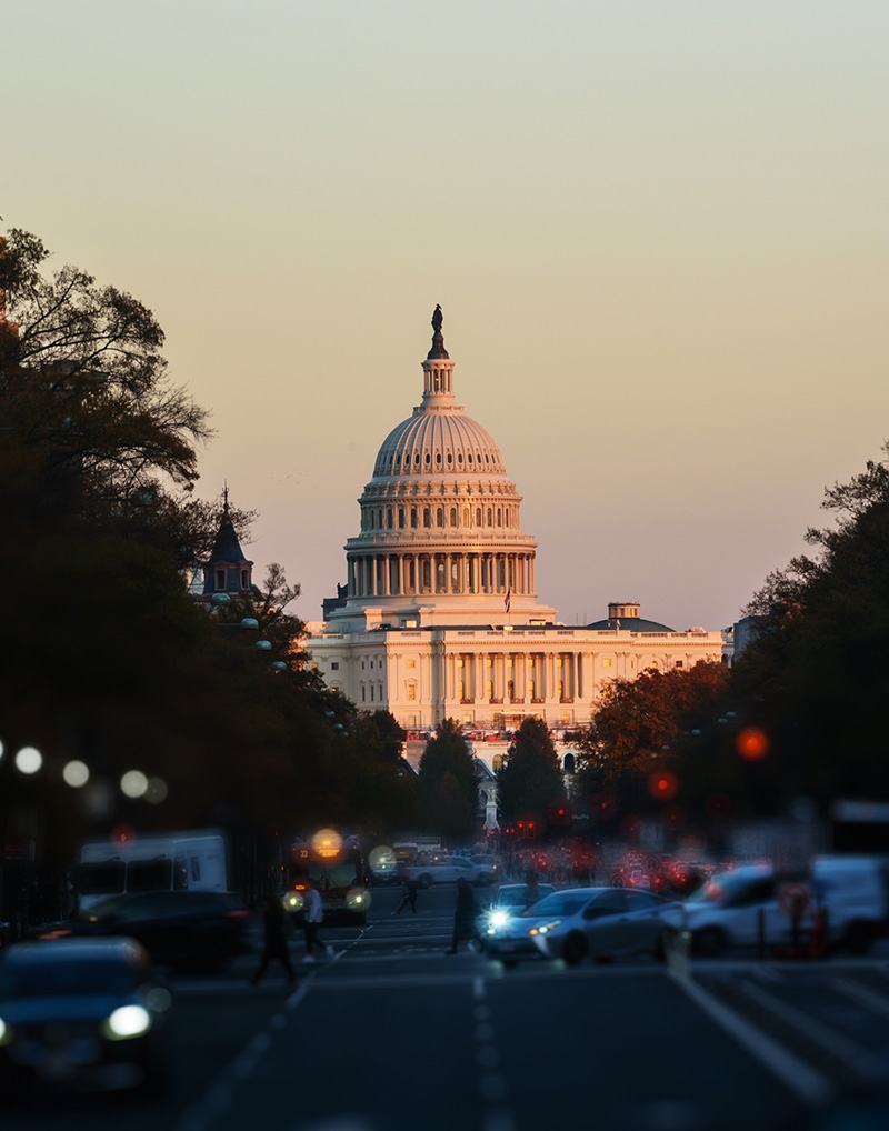 The U.S. Capitol building stands illuminated at sunset, seen from a distance down a busy street with cars and pedestrians in Washington, D.C. Trees frame the scene on both sides.