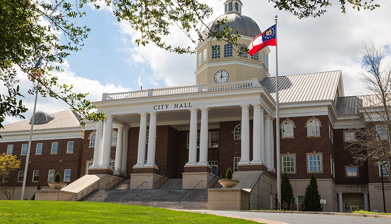 A large brick city hall building with white columns, a central clock tower, and a raised entrance. The Georgia state flag flies on a pole, and trees frame the scene under a partly cloudy sky.