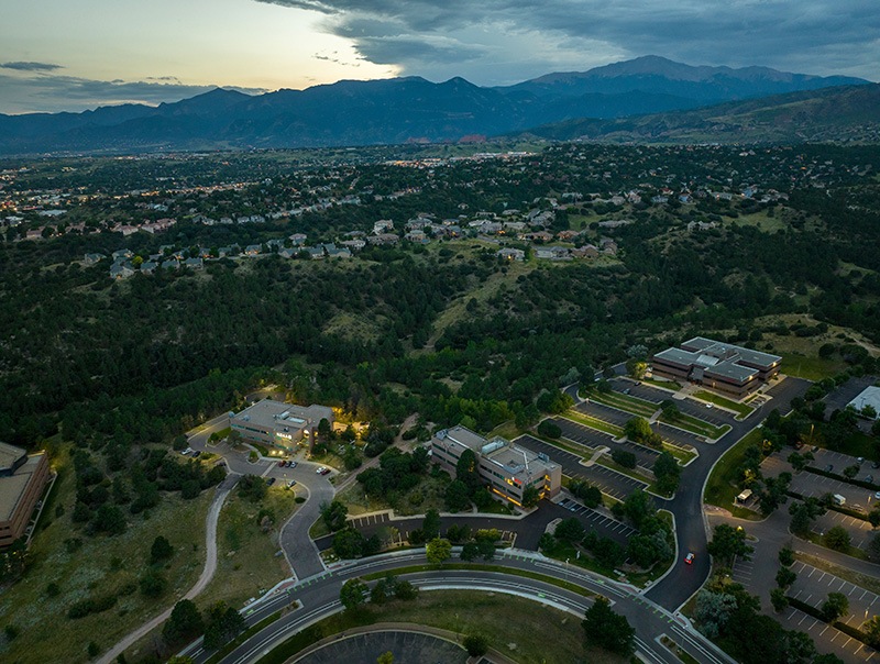Aerial view of a suburban area at dusk, showing office buildings with parking lots, curving roads, dense green trees, and houses scattered across rolling hills, with mountains visible in the distance under a cloudy sky.