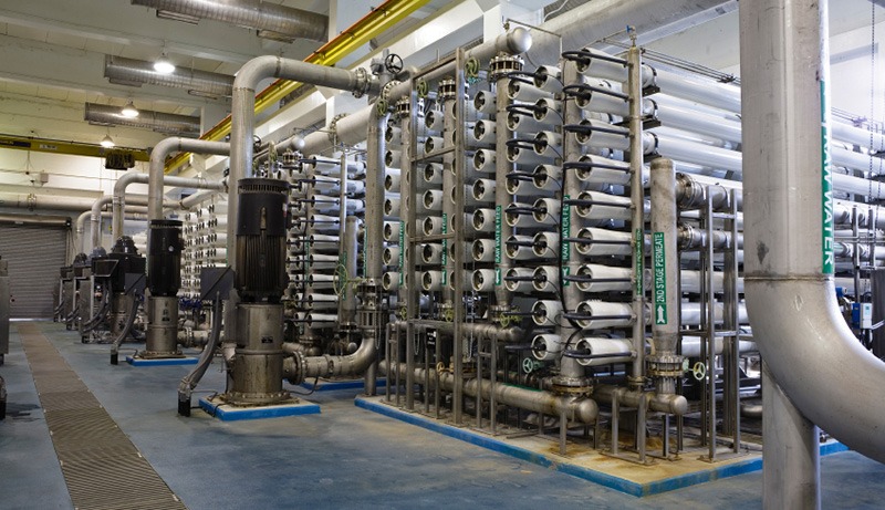 A large industrial facility with rows of cylindrical filtration units, pipes, valves, and pumps, likely used for water treatment or desalination. The area is clean, well-lit, and organized.
