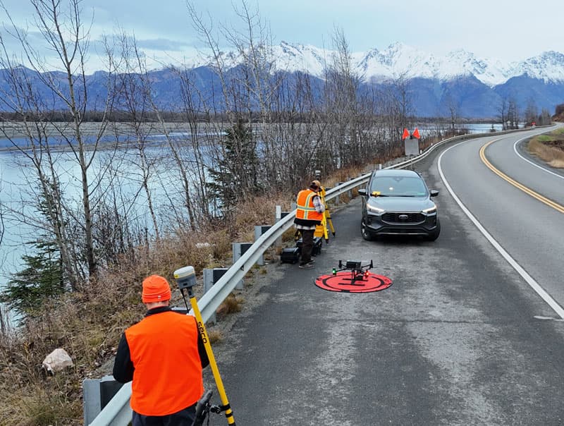 Two workers in orange safety vests operate surveying equipment and a drone on a roadside next to a lake, with a car parked nearby and snow-covered mountains in the background.