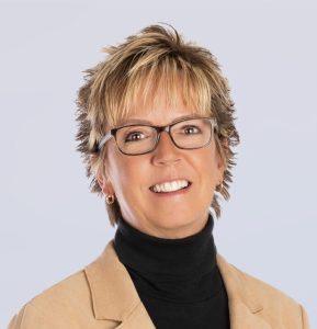 A woman with short, blonde hair and glasses smiles at the camera. She is wearing a beige blazer over a black turtleneck and gold hoop earrings, with a light gray background.