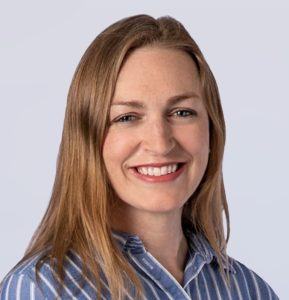 A woman with straight, light brown hair smiles at the camera. She is wearing a blue and white striped collared shirt and is posed against a light grey background.