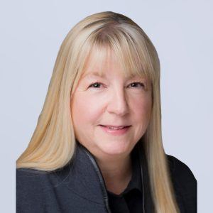 A woman with long, straight blonde hair and bangs is smiling gently. She is wearing a dark jacket and is posed against a plain light background.