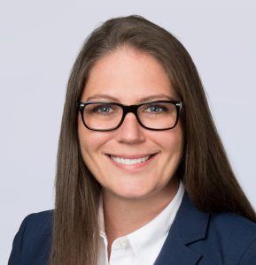 A woman with long brown hair, wearing black glasses, a white collared shirt, and a blue blazer, smiles at the camera against a plain light background.
