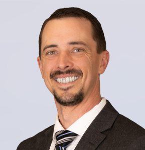 A smiling man with short brown hair and a goatee, wearing a dark suit, white shirt, and striped tie, poses against a light gray background.