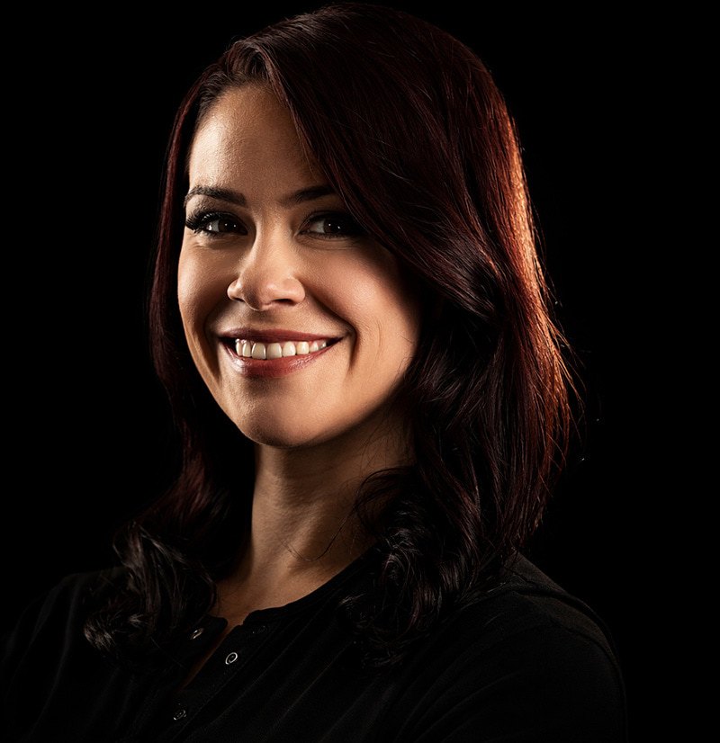 A woman with long, dark hair smiles warmly, wearing a black top against a dark background. The lighting highlights her face and hair, creating a dramatic contrast.